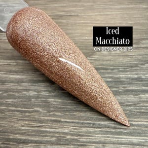 May include: A rose gold glitter nail dip sample in a tapered shape. The sample is labelled "Iced Macchiato" with "CN DESIGNER DIPS" below it. The nail dip has a sparkling, textured appearance. The background is dark.