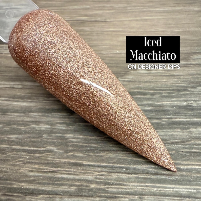 May include: A rose gold glitter nail dip sample in a tapered shape. The sample is labelled "Iced Macchiato" with "CN DESIGNER DIPS" below it. The nail dip has a sparkling, textured appearance. The background is dark.