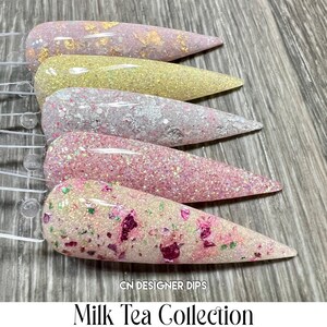 May include: Five nail dip powder colours in a row on a wooden surface. The colours are light pink with gold flakes, yellow, white with silver glitter, pink with silver glitter, and white with pink and green glitter and flakes. The text "CN DESIGNER DIPS" and "Milk Tea Collection" is at the bottom of the image.