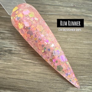 May include: A pink nail swatch with iridescent glitter and hexagonal shapes. The nail is labeled "Rum Runner" by CN Designer Dips. The glitter is a mix of pink, yellow, and purple.