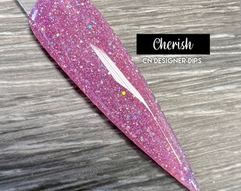 May include: A close-up of a pink nail dip powder with glitter. The powder is in a clear container and has a light pink color with silver glitter. The text "Cherish" and "CN DESIGNER DIPS" is visible in the image.