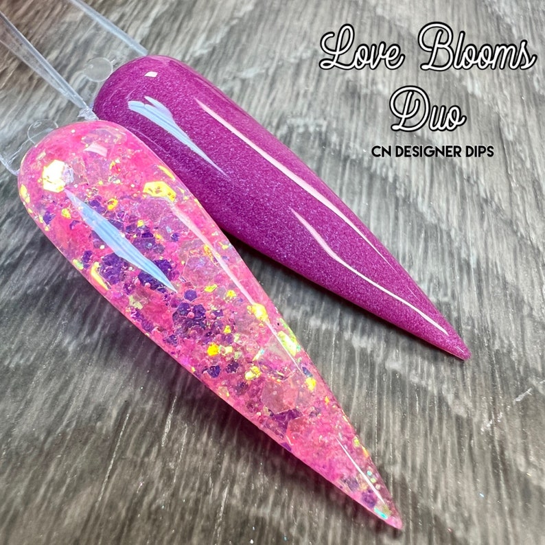 Op de afbeelding: Twee lange, puntige nageltips, een roze met iriserende glitter en een paarse. De tekst "Love Blooms Duo" en "CN DESIGNER DIPS" staat boven de tips.