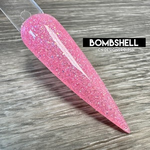 May include: A pink nail dip powder with a glittery finish. The powder is called "Bombshell" and is from CN Designer Dips.