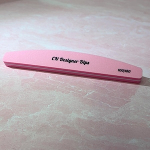 Puede incluir: Una lima de uñas rosa con el texto "CN Designer Dips" y "100/180" impreso. La lima tiene forma rectangular con bordes redondeados y una franja blanca en el lateral.