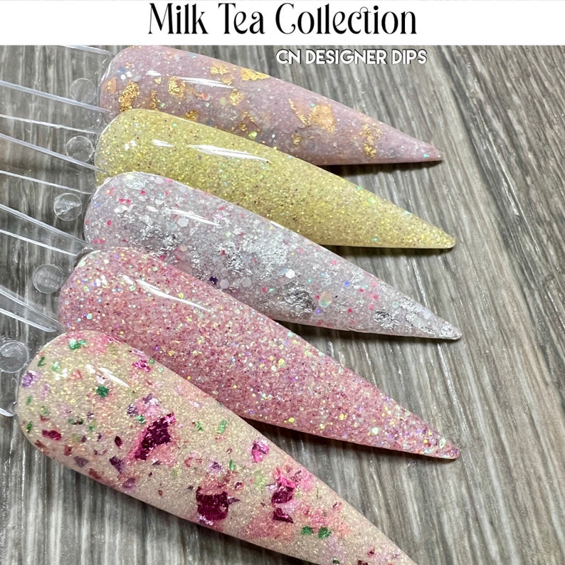 May include: Five different shades of pink, white, and yellow nail polish with glitter and gold flakes. The nail polish is displayed on a clear plastic nail polish holder. The text "Milk Tea Collection" and "CN DESIGNER DIPS" is displayed above the nail polish.
