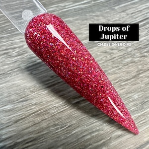 May include: A close-up of a red glitter nail dip powder with a holographic finish. The powder is called "Drops of Jupiter" and is from CN Designer Dips.