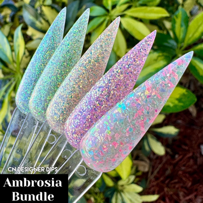 Puede incluir: Cuatro polvos de inmersi&oacute;n para u&ntilde;as transparentes con purpurina iridiscente. Los polvos est&aacute;n dispuestos en fila, con colores que van del azul claro al rosa al blanco. El texto "CN Designer Dips Ambrosia Bundle" es visible en la parte inferior de la imagen.