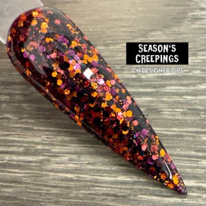 May include: A nail dip sample with a dark base, covered in orange, purple, and red glitter. The text "SEASON'S CREEPINGS" and "CN DESIGNER DIPS" are displayed on a black rectangle.