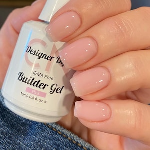 May include: A clear bottle of pink builder gel nail polish with the text "Designer Dreams HEMA Free Builder Gel Pink 15ml-0.5 fl. oz. e" on the label. A hand with pink gel nails is holding the bottle.