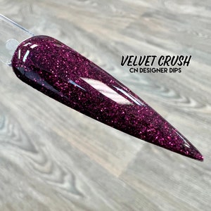 May include: A close-up of a dark purple nail dip powder with a glittery finish. The text "VELVET CRUSH CN DESIGNER DIPS" is visible in the image.