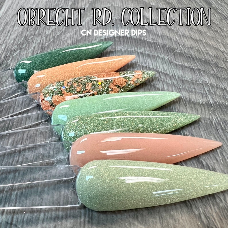 May include: A set of six nail dip powder colors in a variety of green, brown, and beige shades. The colors are arranged in a row on a wooden surface. The text "OBRECHT RD. COLLECTION CN DESIGNER DIPS" is visible at the top of the image.