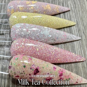 May include: Five different shades of nail dip powder in a milk tea collection. The colours are pink, yellow, white, and two shades of pink with glitter. The dips are displayed on a clear plastic display stand. The text "CN DESIGNER DIPS" and "Milk Tea Collection" is visible on the stand.