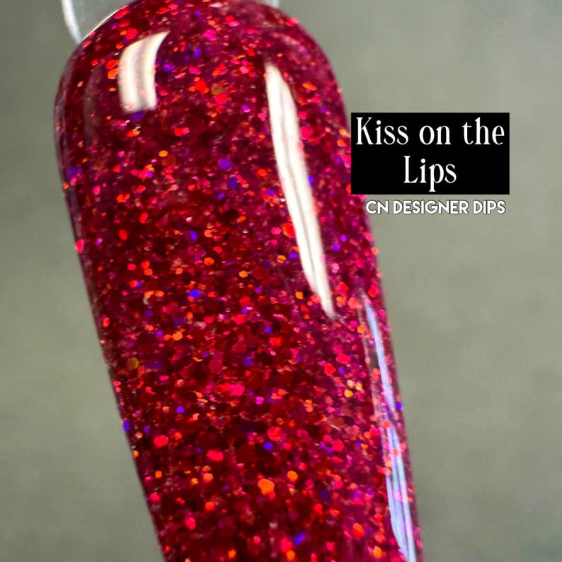 Puede incluir: Primer plano de un esmalte de u&ntilde;as con un tono rojo intenso y brillante. El esmalte est&aacute; salpicado de purpurina roja, naranja y morada de varios tama&ntilde;os. El texto "Kiss on the Lips" y "CN DESIGNER DIPS" es visible.