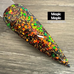May include: A nail swatch with a glitter design. The nail is a dark base with orange, green, and red glitter. The text "Magic Maple" is in a black box, with "CN DESIGNER DIPS" below it.