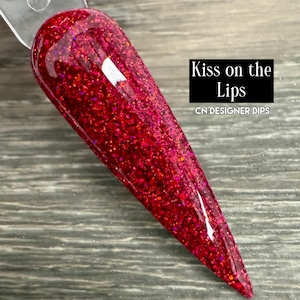 Puede incluir: Una muestra de u&ntilde;as con una vibrante inmersi&oacute;n de purpurina roja. La u&ntilde;a tiene forma de punta. El texto "Kiss on the Lips" y "CN DESIGNER DIPS" se muestran en un rect&aacute;ngulo negro.