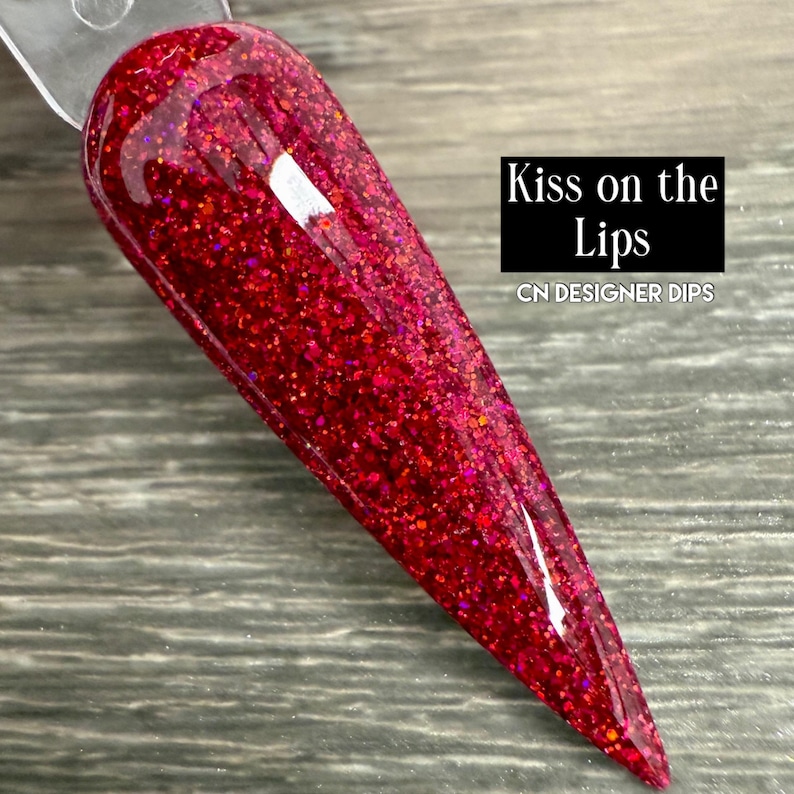 Puede incluir: Una muestra de u&ntilde;as con una vibrante inmersi&oacute;n de purpurina roja. La u&ntilde;a tiene forma de punta. El texto "Kiss on the Lips" y "CN DESIGNER DIPS" se muestran en un rect&aacute;ngulo negro.