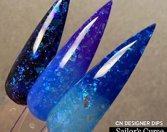May include: Three stiletto-shaped nail swatches displaying the CN DESIGNER DIPS Sailor's Curse Collection. The colours transition from a deep black-blue with iridescent flakes to a vibrant royal blue and a lighter sky blue with metallic accents.