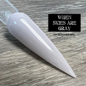 May include: A light gray nail dip powder with a subtle shimmer. The powder is displayed on a clear nail tip. The text "WHEN SKIES ARE GRAY" and "CN DESIGNER DIPS" is visible in the image.