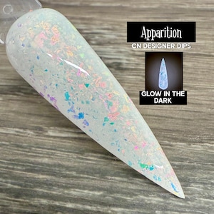 May include: A long, white nail with iridescent glitter and the text "Apparition CN DESIGNER DIPS" and "GLOW IN THE DARK". The nail has a pointed tip and is set against a wooden background. The glitter reflects multiple colors.