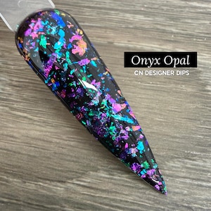 May include: A nail swatch featuring an Onyx Opal design. The nail is black with iridescent flakes in shades of purple, teal, and gold. The text "Onyx Opal" and "CN DESIGNER DIPS" are displayed in a black rectangle.