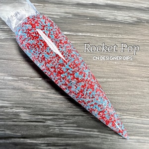 May include: A close-up of a long, pointed nail with a red and blue glitter dip powder. The nail is labeled "Rocket Pop CN DESIGNER DIPS".
