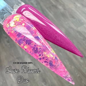 Op de afbeelding: Twee nageldippoederkleuren, &eacute;&eacute;n roze met iriserende glitter en &eacute;&eacute;n dieproze. De tekst "CN DESIGNER DIPS Love Blooms Duo" is onder de nageldips gedrukt.
