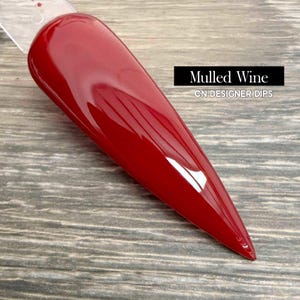 May include: A glossy, elongated nail swatch in a deep red color, labeled "Mulled Wine" by CN Designer Dips. The nail has a pointed tip and a smooth, reflective surface, showcasing the rich hue.
