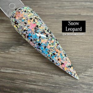 May include: A long, pointed nail sample with a glitter design. The nail features a mix of black, blue, pink, and iridescent glitter, with the text "Snow Leopard" and "CN DESIGNER DIPS" in a black rectangle.