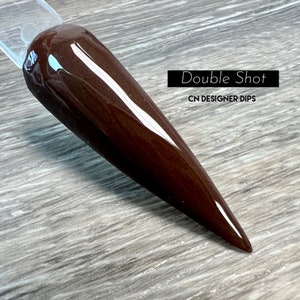 May include: A close-up of a long, pointed, dark brown nail dip. The nail dip is on a wooden surface. The text "Double Shot" and "CN DESIGNER DIPS" is visible in the image.