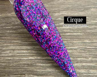 May include: A close-up of a long, pointed nail with a glittery, multi-colored dip powder. The colors are predominantly pink and blue with a hint of purple. The nail is on a wooden surface. The text "Cirque CN DESIGNER DIPS" is visible in the image.