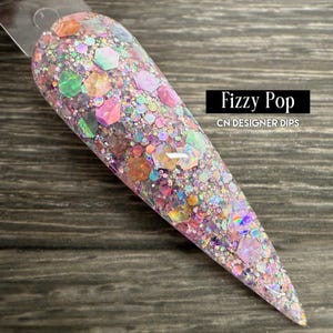 May include: A nail swatch with a glittery, multi-colored design. The design features a mix of iridescent and opaque glitter in various shapes and sizes, including large hexagonal pieces. The text "Fizzy Pop" and "CN DESIGNER DIPS" is visible.