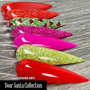 May include: Six different colors of glitter nail dip powder displayed on nail tips. The colors are red, green, pink, lime green, and red. The text "CN DESIGNER DIPS Dear Santa Collection" is at the bottom of the image.