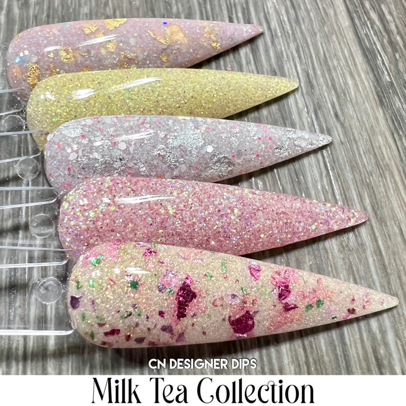 May include: Five different shades of nail dip powder in a milk tea collection. The colours are a light pink with gold flakes, a light yellow with glitter, a white with glitter, a light pink with glitter, and a white with pink and green glitter.