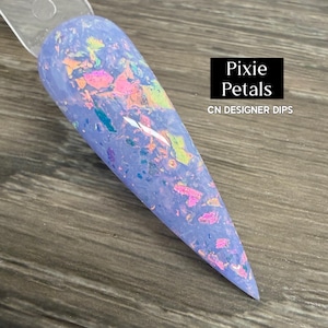 May include: A light purple nail with iridescent flakes. The nail is shaped like a long almond and is held by a clear plastic stick. The text "Pixie Petals" and "CN DESIGNER DIPS" are in a black rectangle.