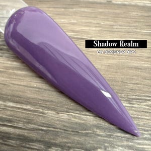 May include: A long, purple nail tip with a glossy finish. The nail is a solid color, with the text "Shadow Realm" and "CN DESIGNER DIPS" in a black rectangle.