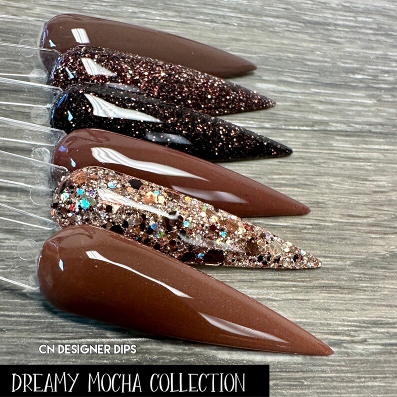 May include: A set of five brown nail dip powders with glitter. The powders are arranged in a row on a wooden surface. The text "CN DESIGNER DIPS DREAMY MOCHA COLLECTION" is visible at the bottom of the image.