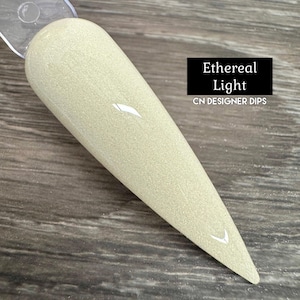 May include: A close-up of a light beige nail dip powder with a shimmery finish. The powder is in a clear plastic container and is labeled "Ethereal Light CN DESIGNER DIPS".