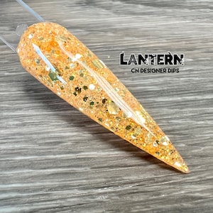 May include: A close-up of a long, pointed nail with a peach-colored base and gold glitter. The nail is on a wooden surface. The text "LANTERN CN DESIGNER DIPS" is visible in the upper right corner of the image.