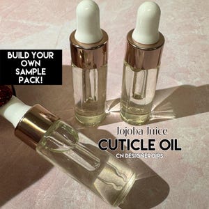 Puede incluir: Tres botellas de vidrio transparente de aceite para cutículas con cuentagotas blancos y detalles en oro rosa. Las botellas están etiquetadas como "Jojoba Juice CUTICLE OIL" y "CN DESIGNER DIPS". Un rectángulo negro dice "BUILD YOUR OWN SAMPLE PACK!"