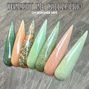 May include: A set of seven nail dip powder colors in a variety of green and peach shades. The colors are arranged in a row on a clear display stand. The text "OBRECHT RD. COLLECTION CN DESIGNER DIPS" is visible at the top of the image.