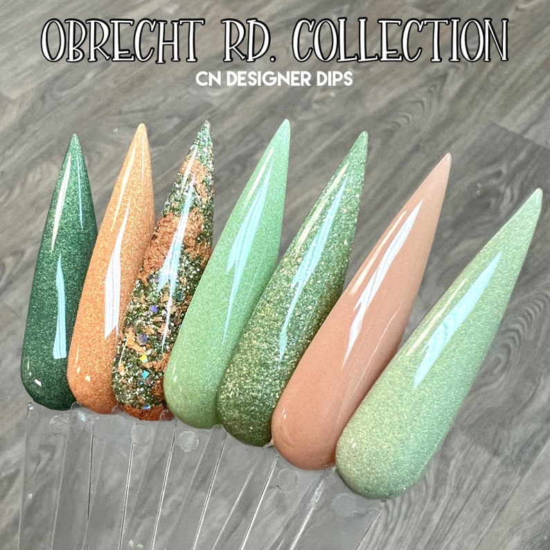 May include: A set of seven nail dip powder colors in a variety of green and peach shades. The colors are arranged in a row on a clear display stand. The text "OBRECHT RD. COLLECTION CN DESIGNER DIPS" is visible at the top of the image.