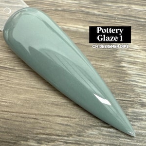 May include: A long, pointed nail sample in a muted green-gray color. The sample is labeled "Pottery Glaze 1" with "CN DESIGNER DIPS" below it in a black rectangle. The nail has a glossy finish and is set against a wooden surface.