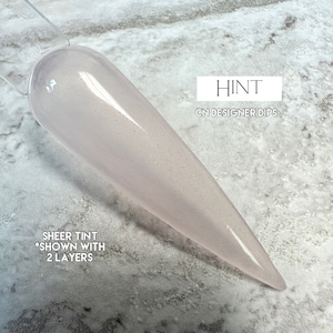 May include: A light pink nail dip powder with a sheer tint. The text "HINT" and "CN DESIGNER DIPS" is visible on the label. The text "SHEER TINT *SHOWN WITH 2 LAYERS" is also visible on the label.