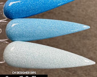May include: Three stiletto-shaped nail swatches featuring a gradient of blue glitter finishes, ranging from a deep sky blue at the top to a pale icy blue at the bottom. The text CN DESIGNER DIPS Smooth Sailing Tonal is printed below.