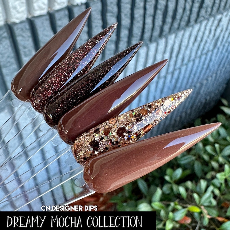 May include: Five different shades of brown nail dip powder, including a glitter shade. The text "CN DESIGNER DIPS DREAMY MOCHA COLLECTION" is at the bottom of the image.