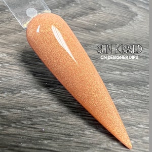 May include: A close-up of a long, pointed, light orange nail polish with a shimmery finish. The nail polish is on a wooden surface. The text "SUN KISSED CN DESIGNER DIPS" is visible in the image.
