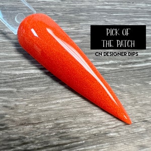 May include: A close-up of a bright orange nail dip powder with a glittery finish. The text "PICK OF THE PATCH CN DESIGNER DIPS" is visible in the background.