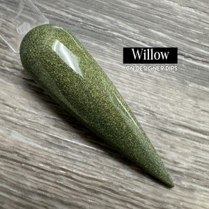May include: A nail swatch displays a glittery, olive-green nail dip powder. The text "Willow" is in a black rectangle, with "CN DESIGNER DIPS" below it. The nail is long and pointed, with a clear plastic handle.