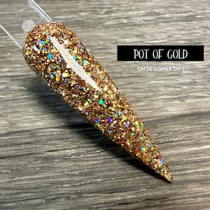 May include: A nail swatch with a glittery, gold dip powder design. The nail is shaped with a pointed tip and features the text "POT OF GOLD" and "CN DESIGNER DIPS" in a black rectangle.