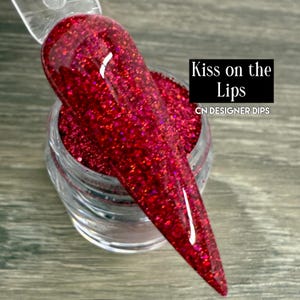 Puede incluir: Primer plano de una u&ntilde;a sumergida en purpurina roja vibrante. La u&ntilde;a se sostiene sobre un peque&ntilde;o tarro transparente lleno de la misma purpurina. El texto "Kiss on the Lips" y "CN DESIGNER DIPS" son visibles.